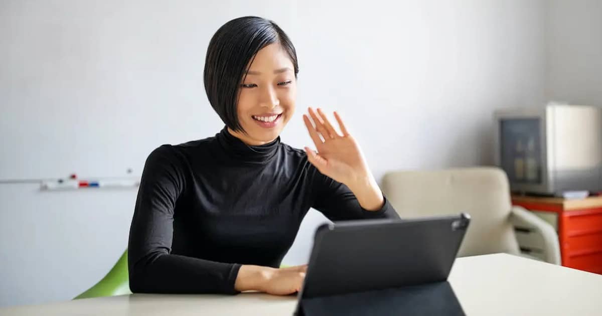 Asian woman smiling and waving while video chatting via tablet