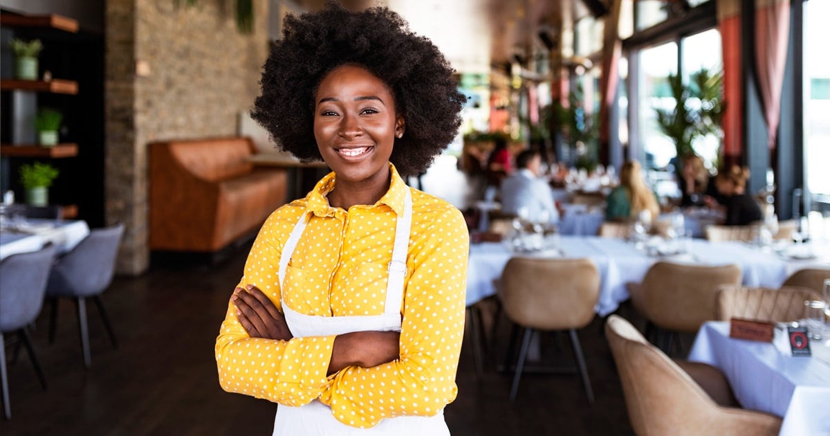 Black female coffee shop owner with natural hair smiling while wearing wearing apron in the dining area