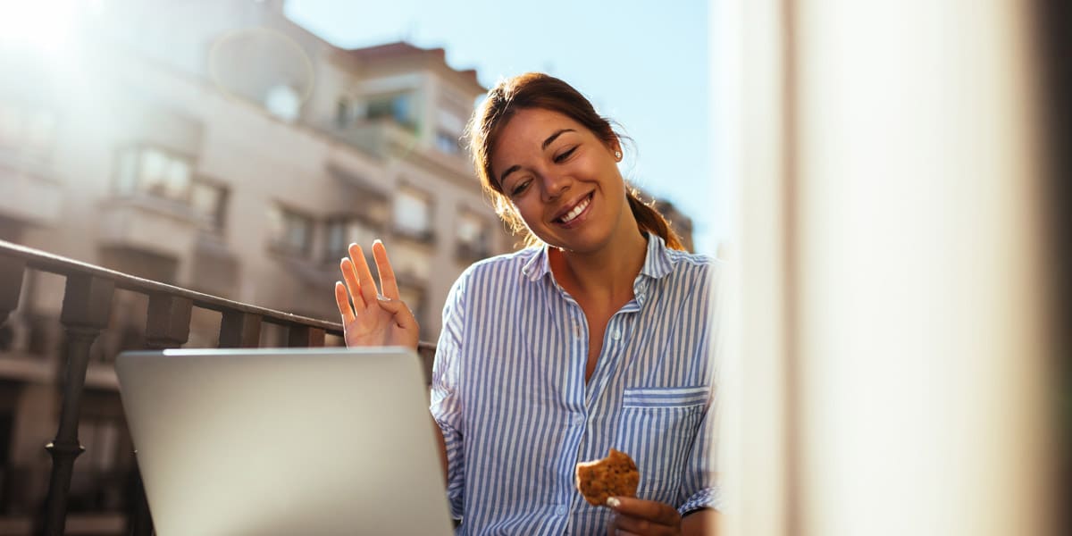 a cheerful young woman staying connected and chatting via laptop while enjoying a bite to eat on the balcony